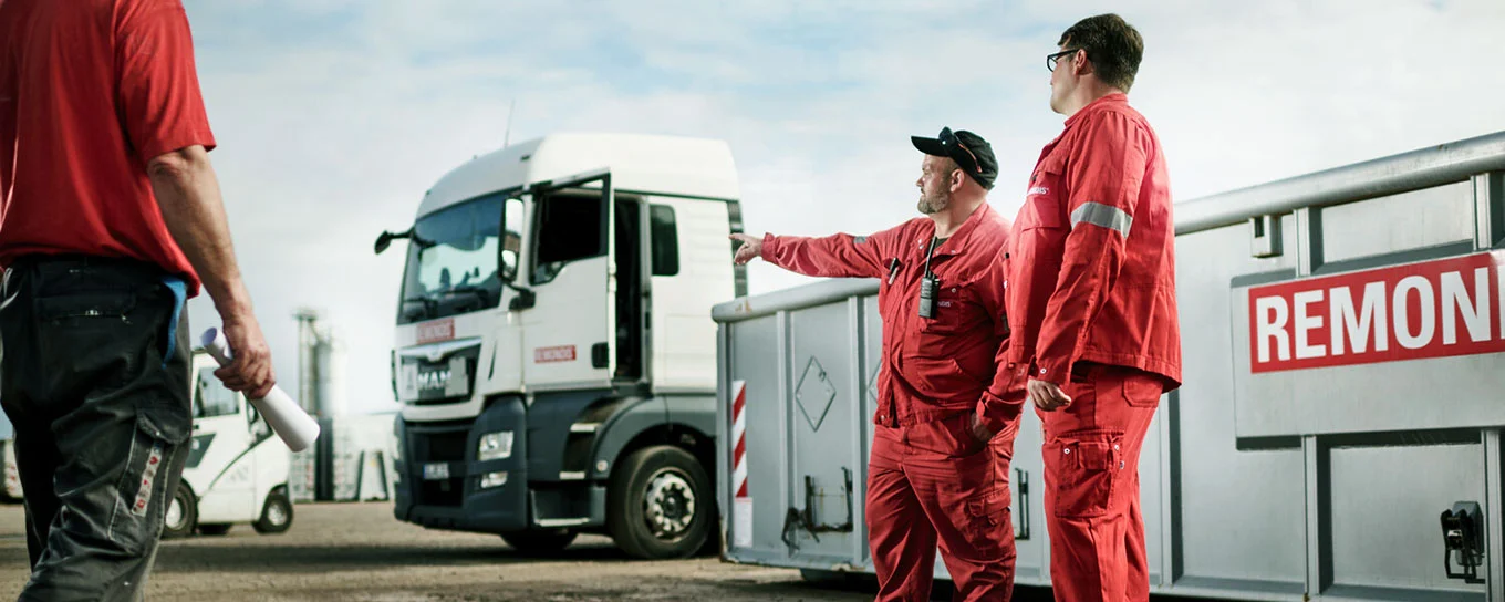 Tre dipenti di REMONDIS in tenuta da lavoro in un centro di riciclaggio parlano davanti a un grande cassone e un camion.