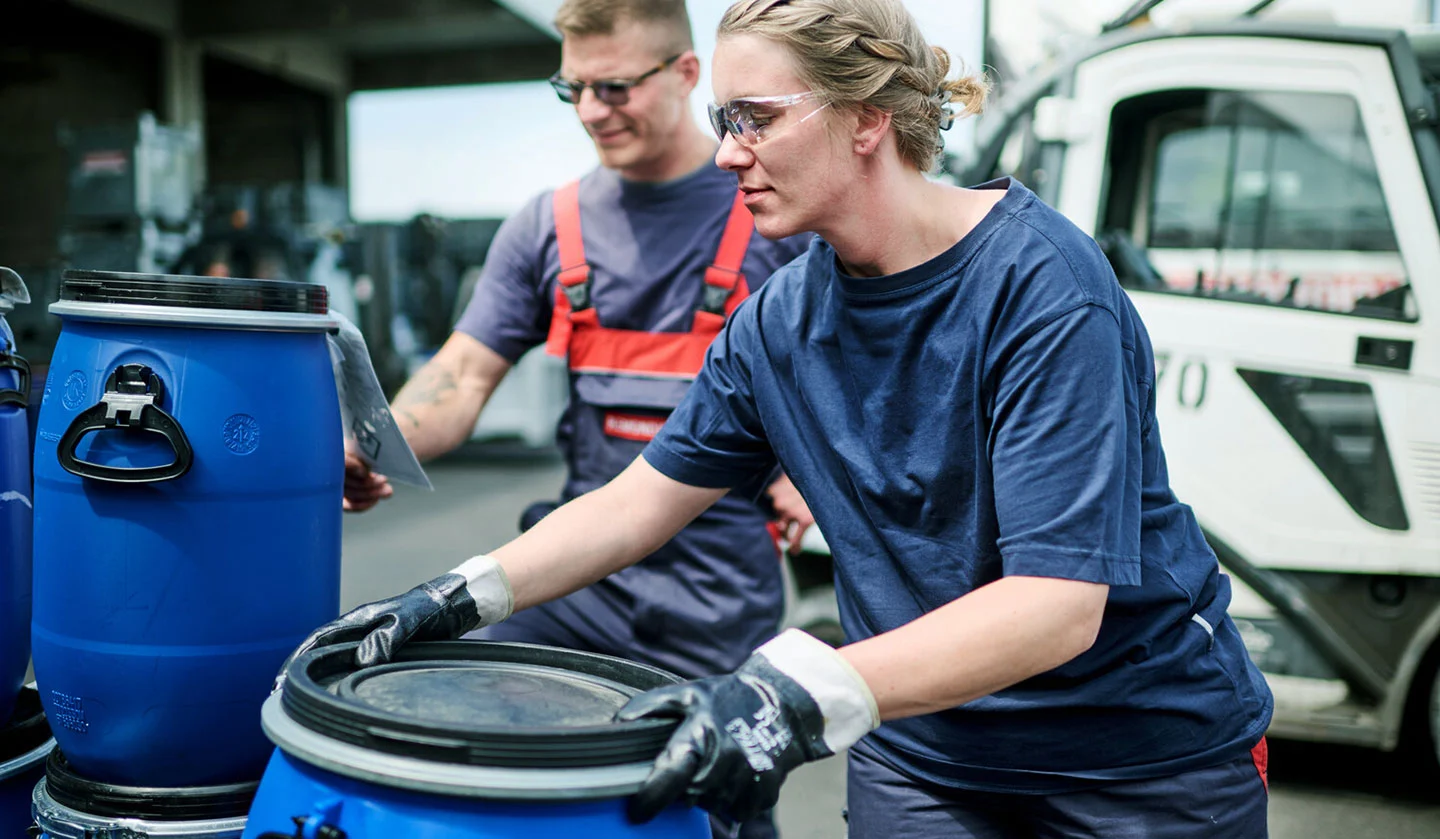 Due dipendenti maneggiano fusti blu con anello di tenuta; sullo sfondo un carrello elevatore